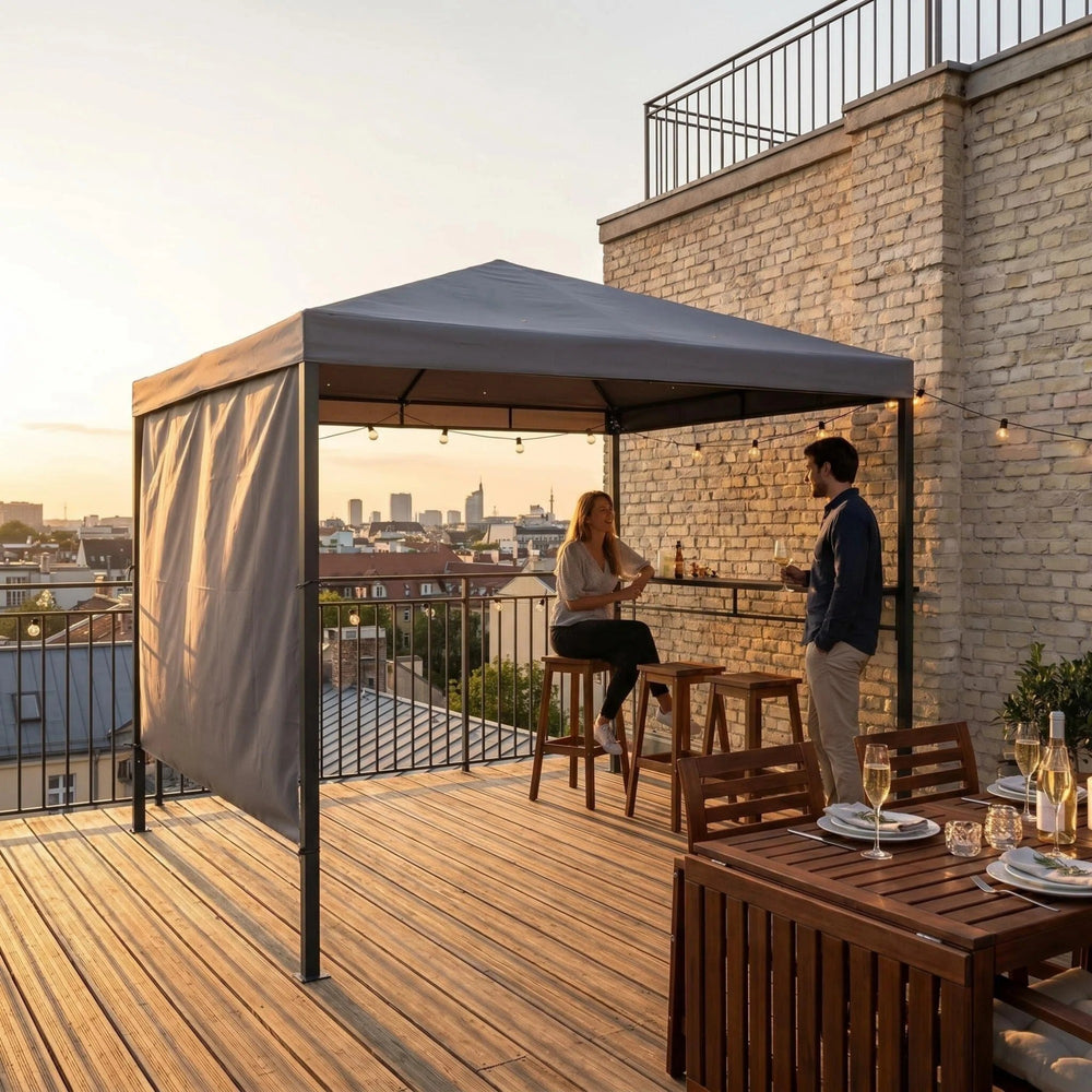 A gray outdoor patio canopy with side curtains is set up on a wooden rooftop terrace at sunset, with two people standing and sitting underneath near a brick wall and dining table.